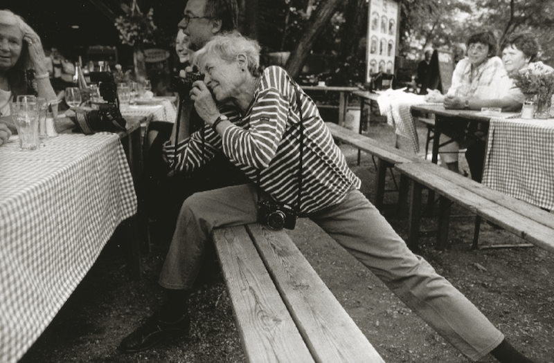 Black and white photograph of Inge Morath stretching across a bench to capture a photograph of a couple seated